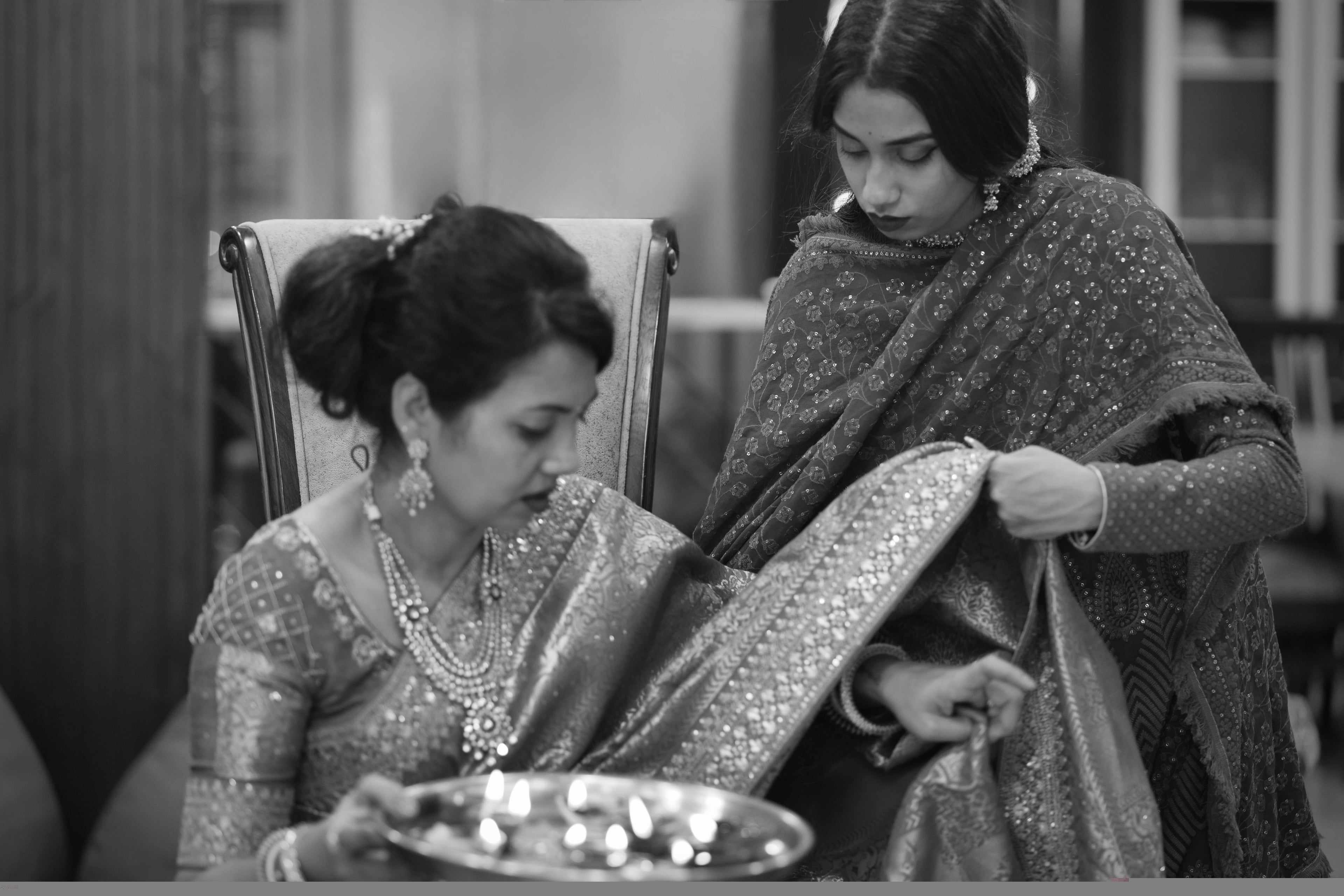 Two women in traditional attire sitting together, one adjusting the other's shawl.